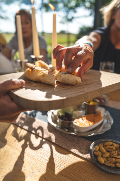 Verschiedene Tapas auf dem Tisch, ein Gast reicht ein Holzbrett mit Tapas dem Gegen&uuml;ber, w&auml;hrend die Sonne in den Gastraum scheint.