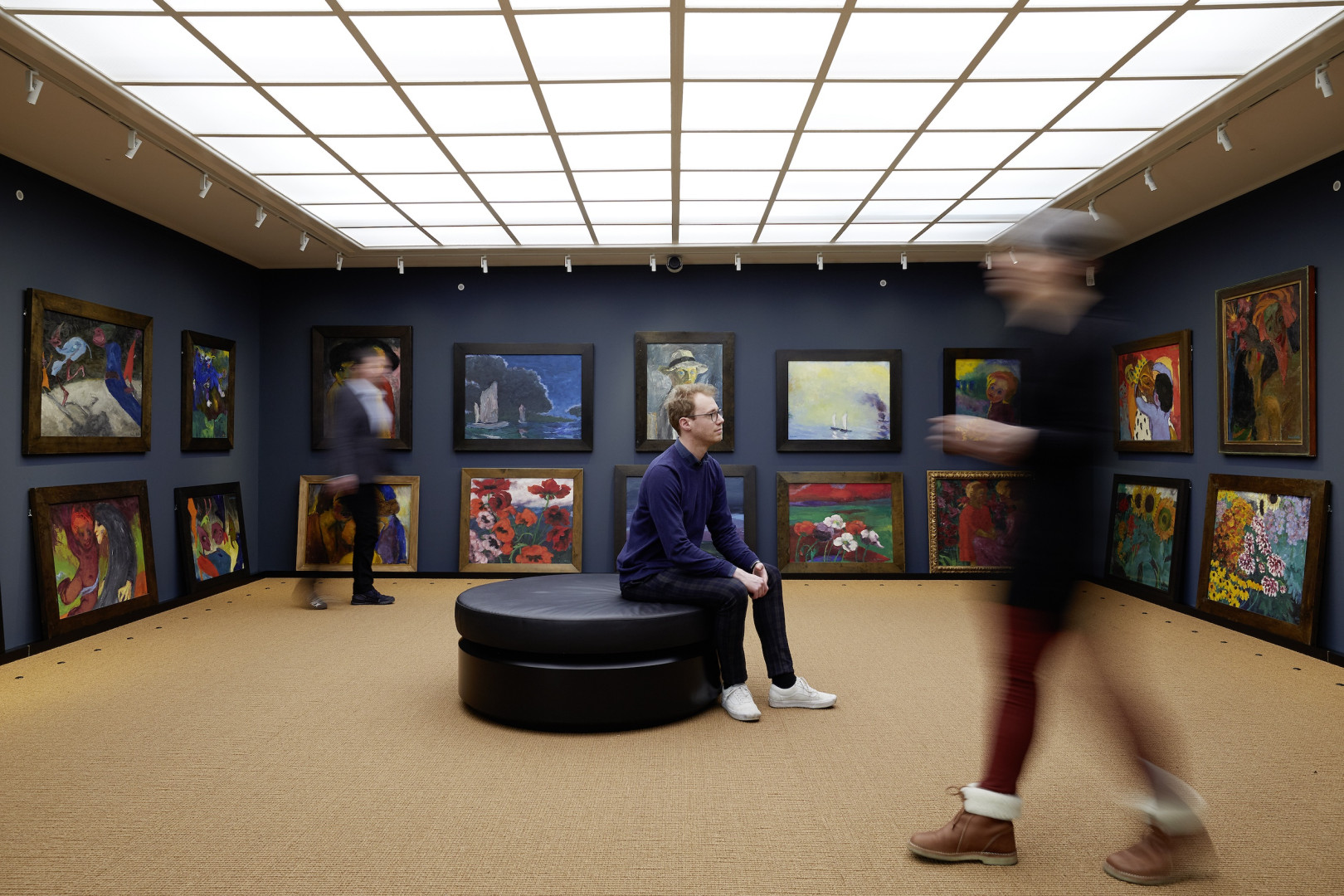 Besucherinnen und Besucher im Bildersaal des Nolde-Hauses vor dunkelblauen Wänden mit dicht gehängten Gemälden von Emil Nolde.