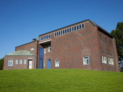 Au&szlig;enansicht des Nolde-Hauses mit blauer Eingangst&uuml;r, rotem Klinker und markanter Fensterfront des Bildersaals; der Bau erinnert an den Bauhausstil der 1920er Jahre.